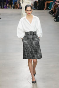 Model on runway wearing a white oversized blouse with puffed sleeves, tucked into a high-waisted black-and-white plaid belted skirt, with matching plaid heels.