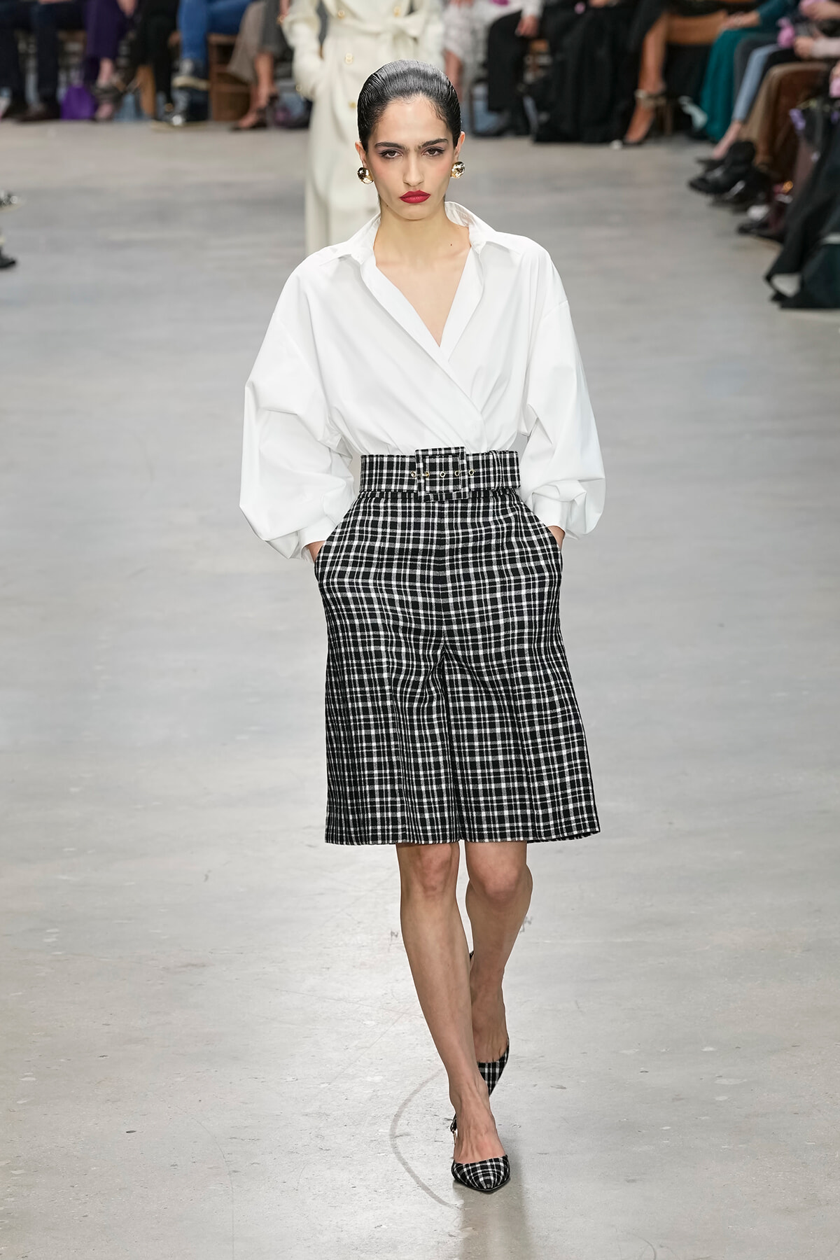 Model on runway wearing a white oversized blouse with puffed sleeves, tucked into a high-waisted black-and-white plaid belted skirt, with matching plaid heels.