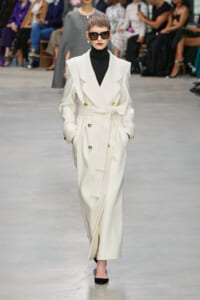 Model walking a fashion show in a white belted double-breasted trench coat with gold buttons, black turtleneck, and oversized sunglasses; audience blurred in background.