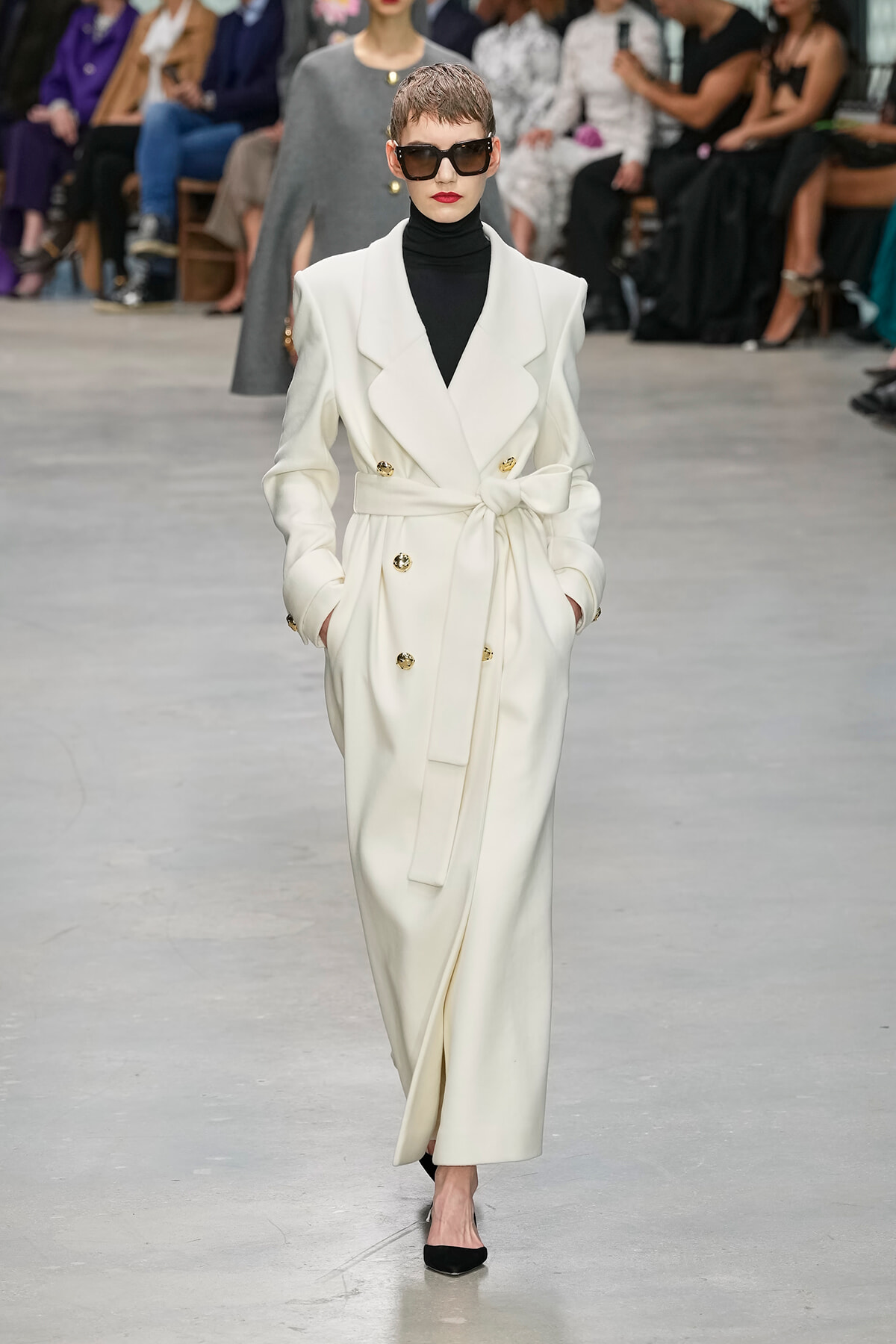 Model walking a fashion show in a white belted double-breasted trench coat with gold buttons, black turtleneck, and oversized sunglasses; audience blurred in background.