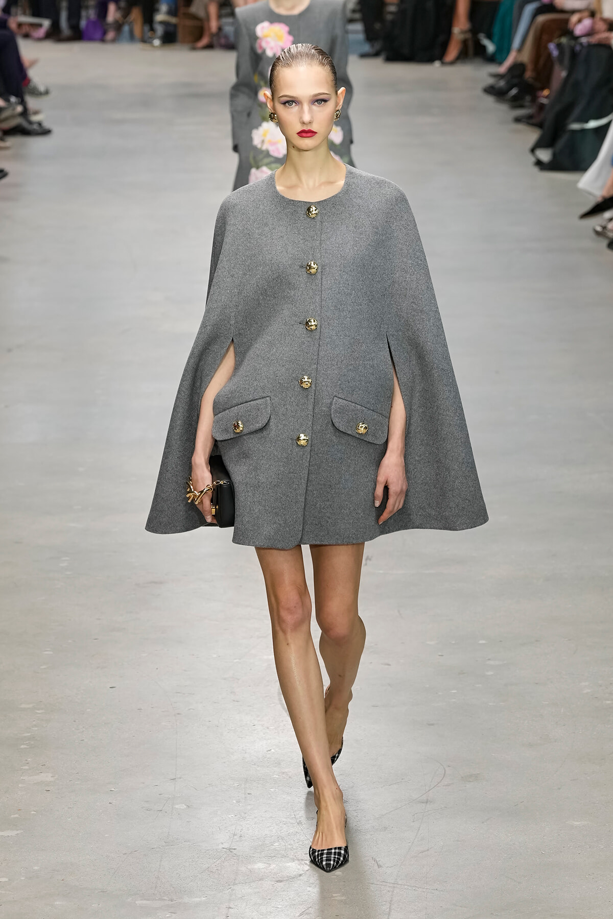 Model walks the runway wearing a gray cape-style coat with gold buttons and oversized front pockets, holding a small black clutch.