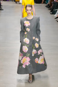 Model walks a runway in a long gray dress adorned with large pink and yellow floral embroidery; she has slicked-back hair and pink floral heels.