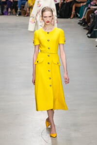 Model walks the runway in a bright yellow, textured button-front dress with belt and pocket flaps, paired with yellow heels.