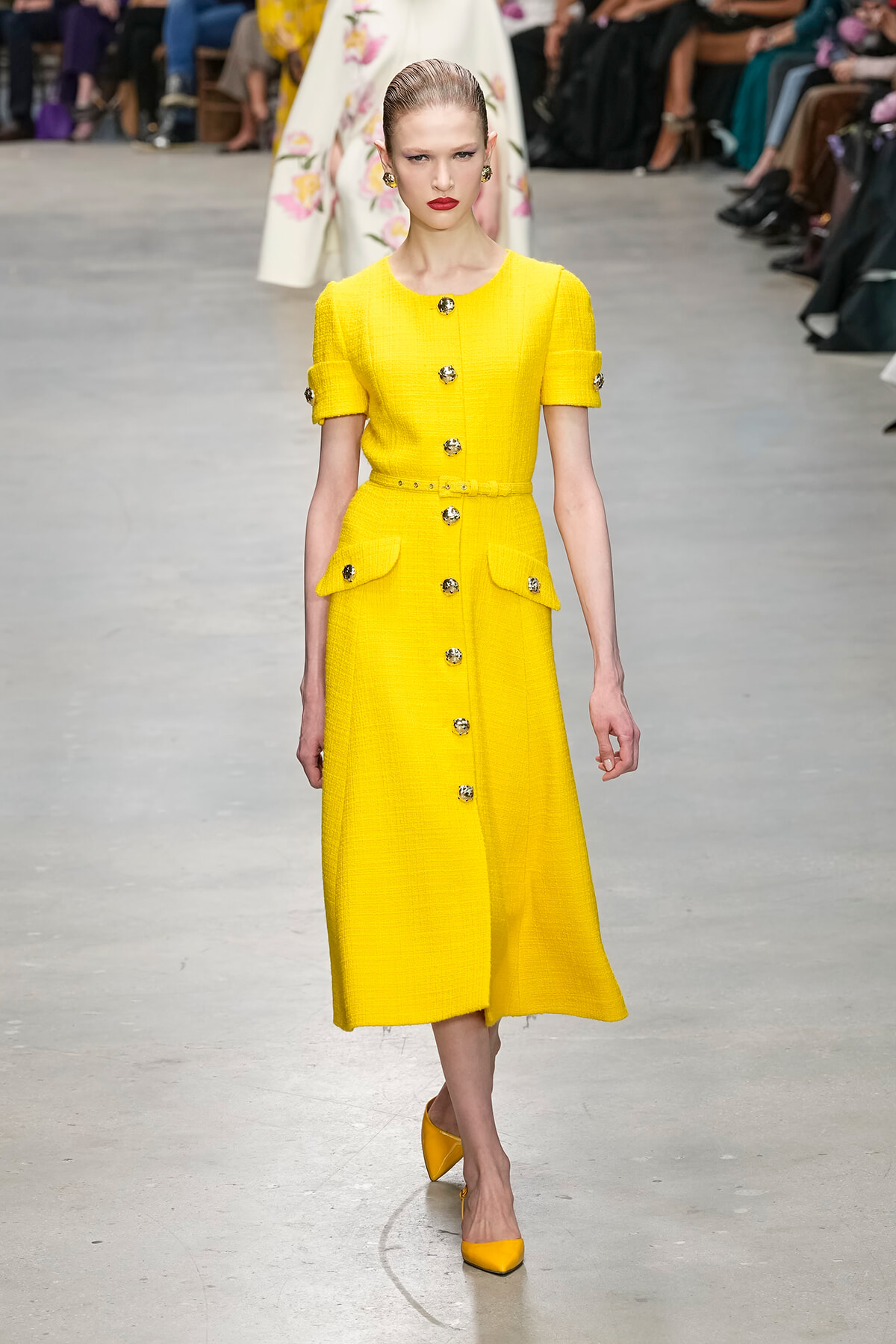 Model walks the runway in a bright yellow, textured button-front dress with belt and pocket flaps, paired with yellow heels.