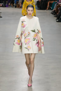 Model walks a fashion runway in a white cape coat with pink and yellow floral embroidery, paired with pink heels.