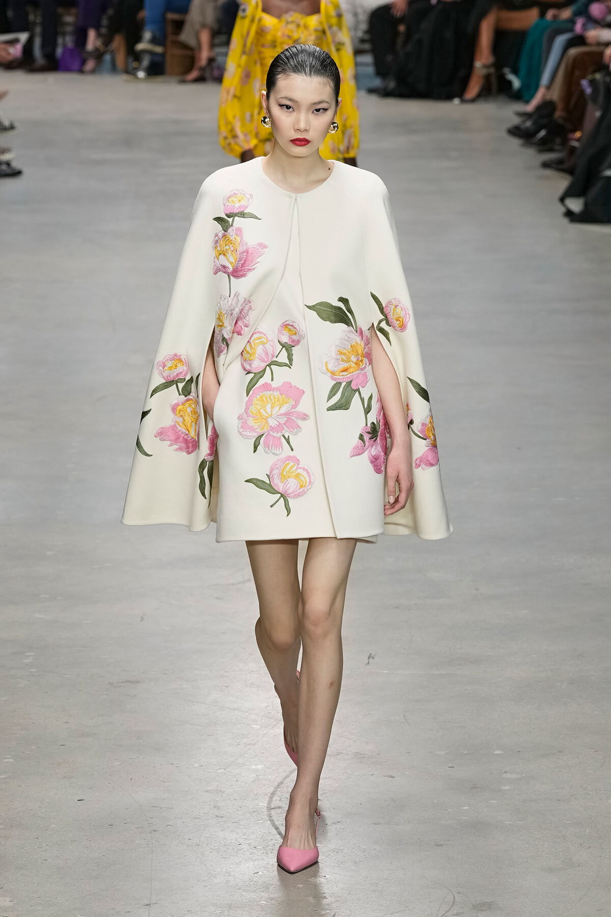 Model walks a fashion runway in a white cape coat with pink and yellow floral embroidery, paired with pink heels.