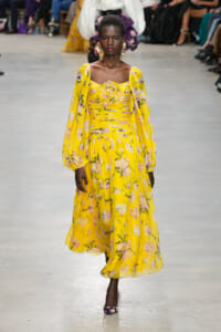 Model wearing a bright yellow floral gown with off-shoulder bodice and puff sleeves walking a runway.