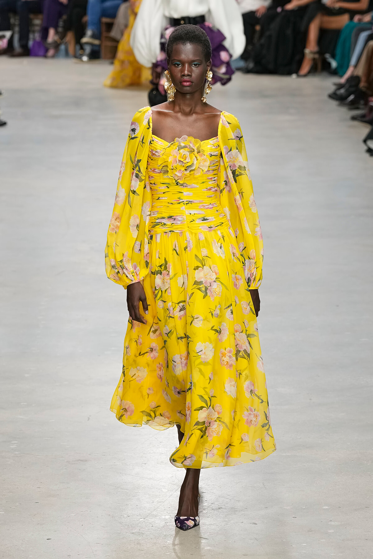 Model wearing a bright yellow floral gown with off-shoulder bodice and puff sleeves walking a runway.