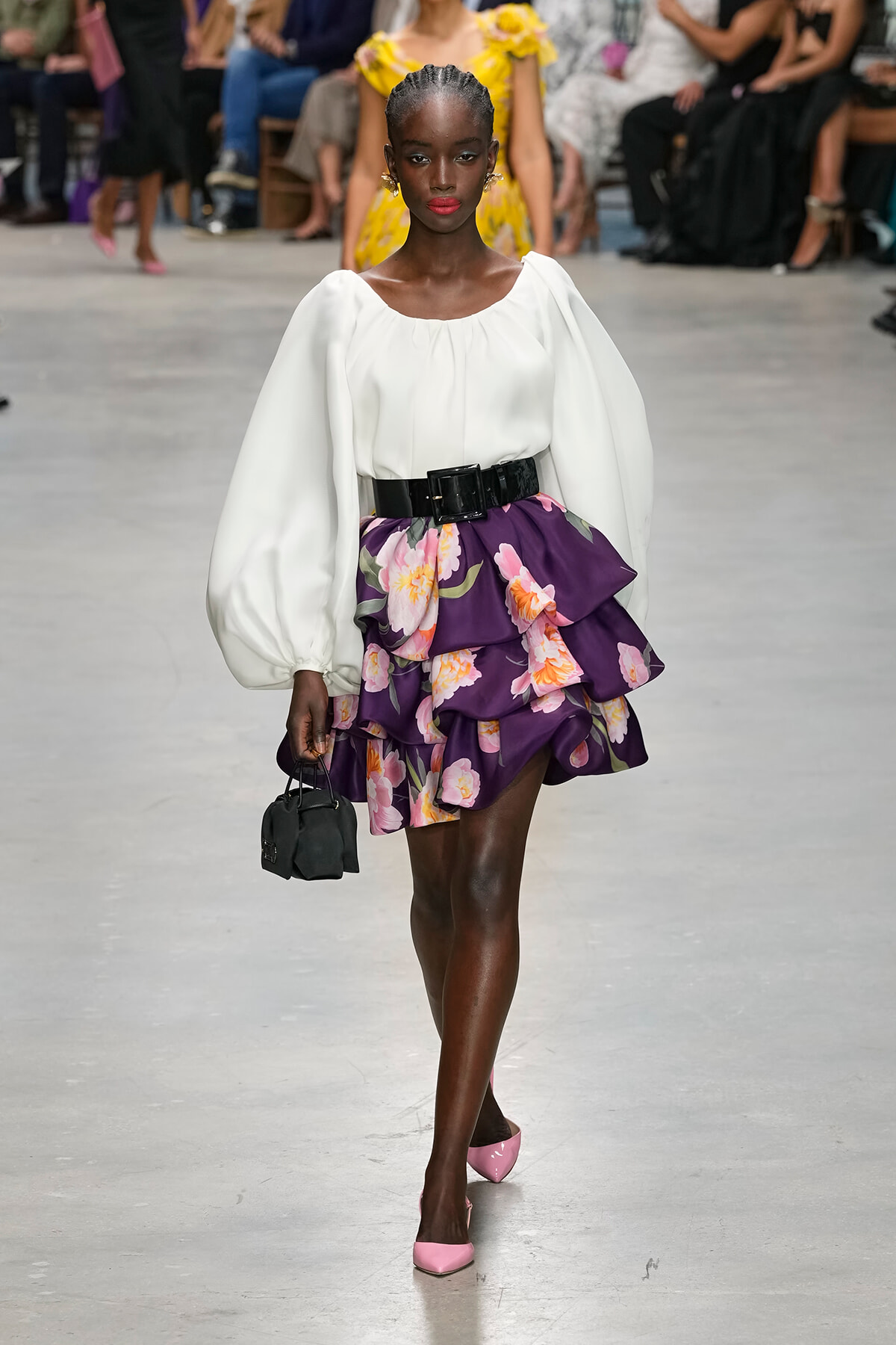 Model walks a runway in a white puff-sleeve blouse and a purple floral layered skirt with a wide black belt, pink heels, and a small black handbag.