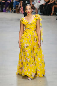 Model walks a fashion runway in a bright yellow floral maxi dress with an off‑the‑shoulder draped bodice and a large flower adornment at the shoulder.
