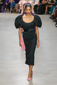 Model walks a fashion runway in a black puff-sleeve dress with a wide belt, pink clutch, and pink heels amid a blurred audience.