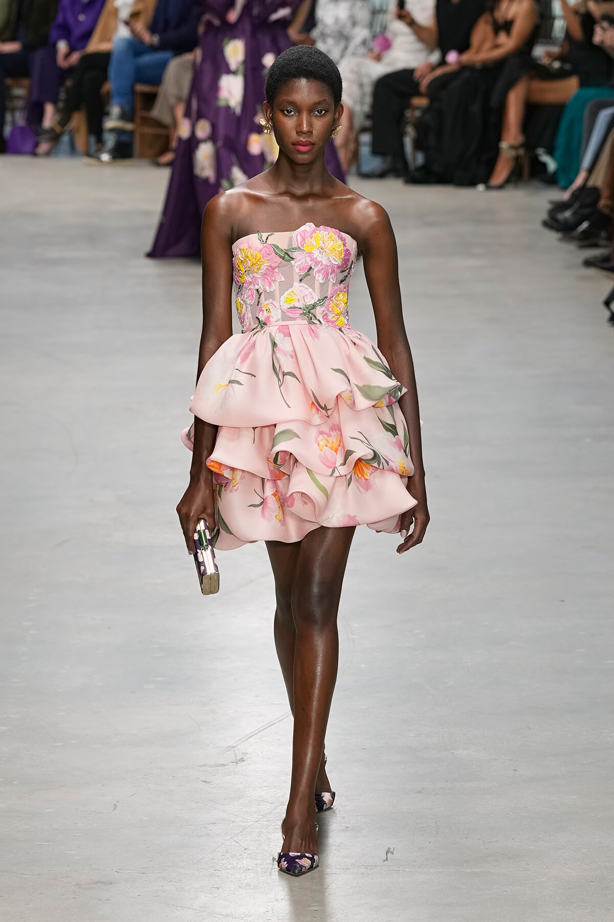 Female model walks the runway in a pink strapless floral dress with layered ruffles, holding a small clutch.