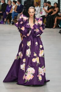 Model walking the runway in a floor-length purple floral gown with large ruffled shoulders and pink-white flowers, black earrings, and a slicked-back hairstyle.