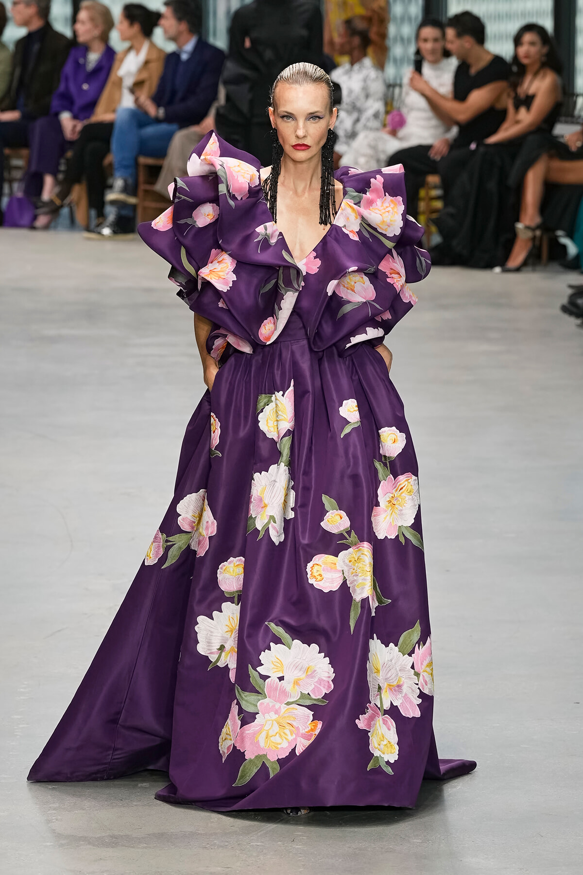Model walking the runway in a floor-length purple floral gown with large ruffled shoulders and pink-white flowers, black earrings, and a slicked-back hairstyle.