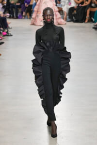Model walks the runway in a black turtleneck dress with a dramatic oversized ruffled train and long beaded earrings during a fashion show.