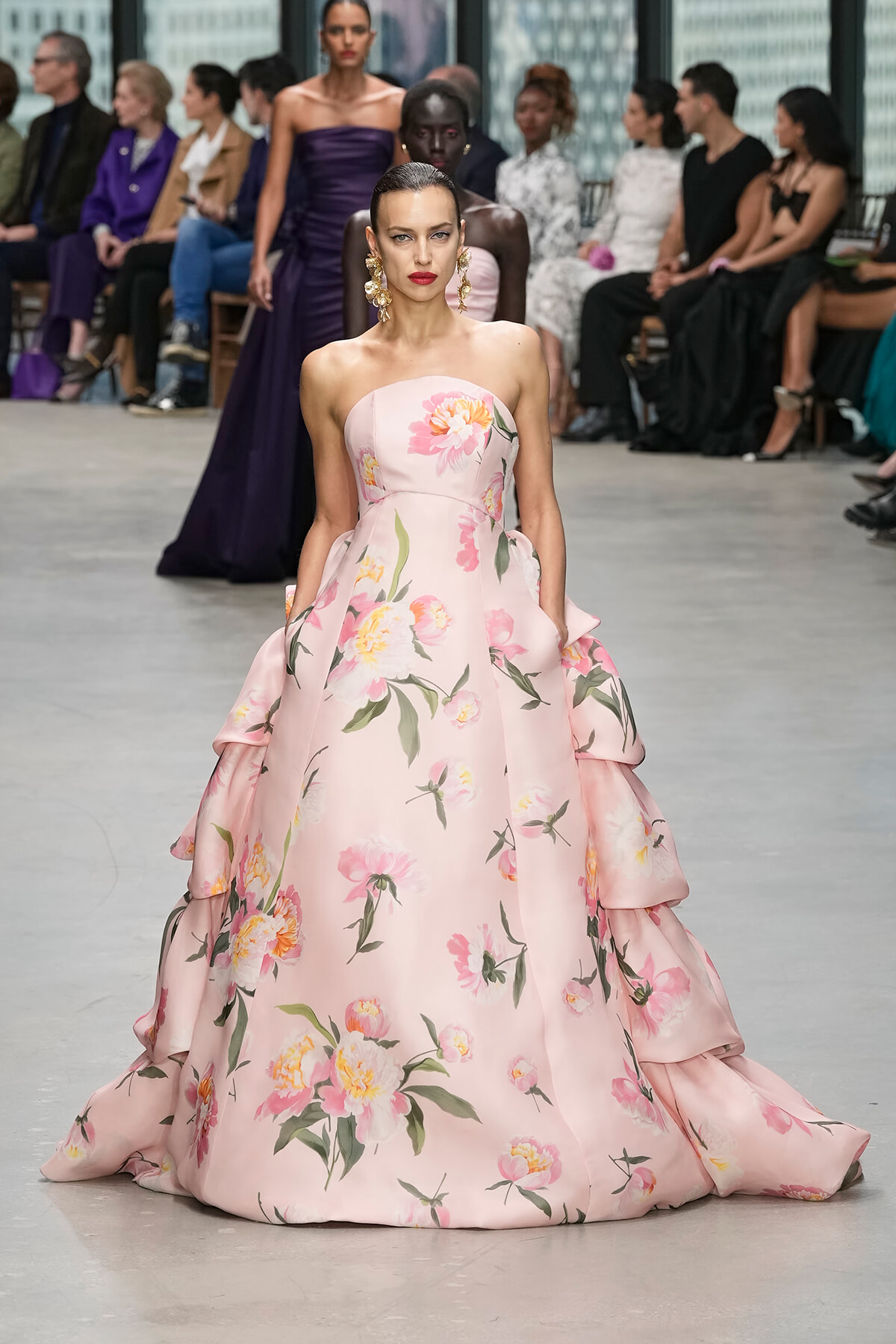 Model walks the runway in a pale pink, voluminous floral gown with large yellow-pink blossoms and green leaves, wearing bold gold statement earrings.