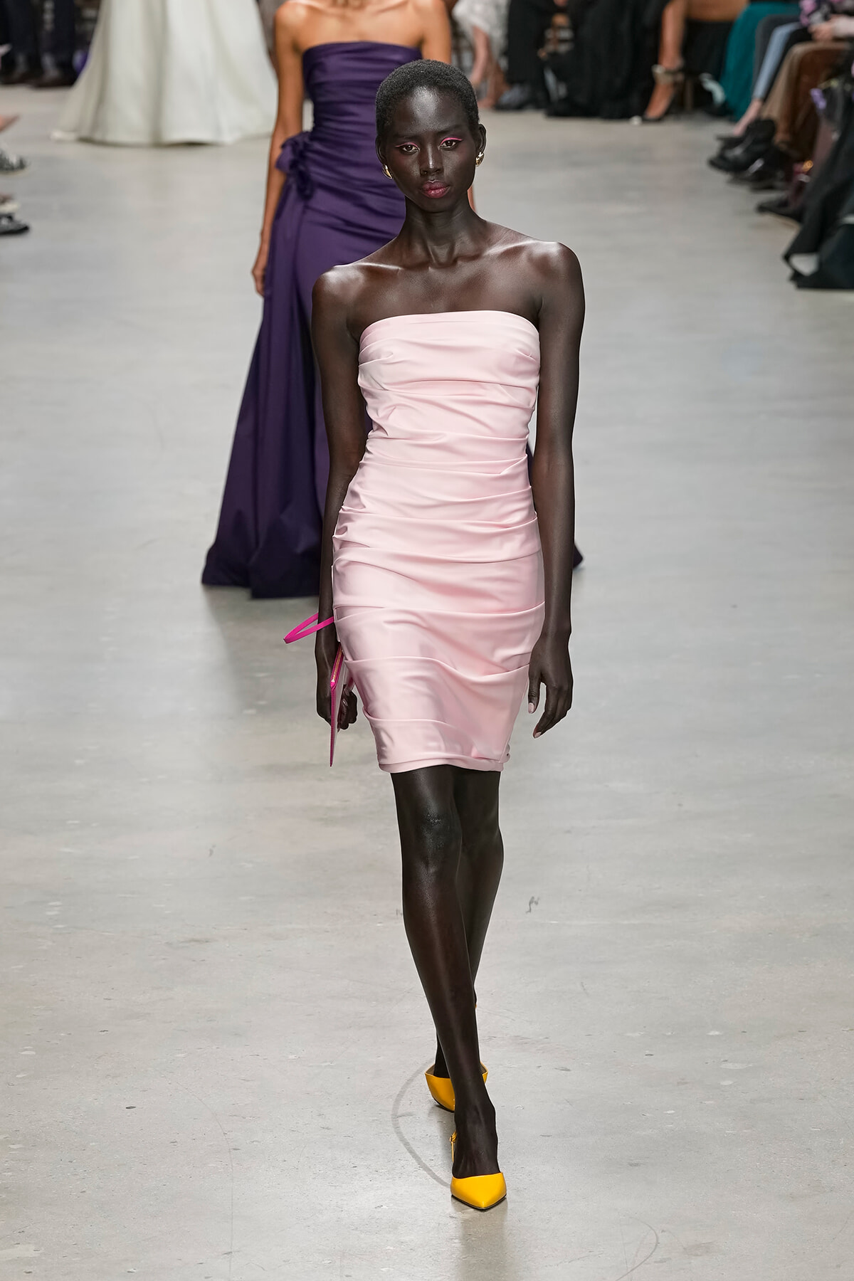 Black model walks a fashion show wearing a pale pink strapless ruched mini dress and bright yellow heels with a purple gown visible in the background.