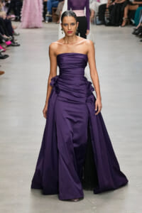 Model walks runway wearing a strapless, deep purple satin gown with gathered bodice and ruffled side detailing.