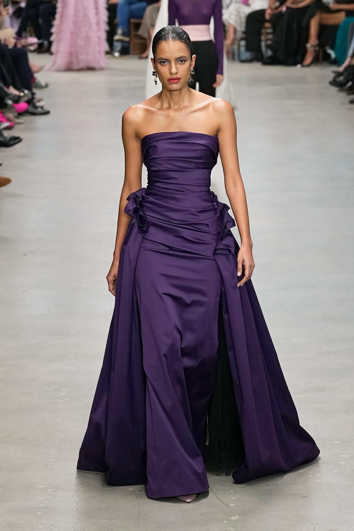 Model walks runway wearing a strapless, deep purple satin gown with gathered bodice and ruffled side detailing.
