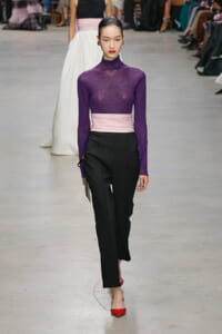 Model walking a fashion runway in a purple sheer turtleneck, pale pink sash belt, black trousers, and red pointed flats.