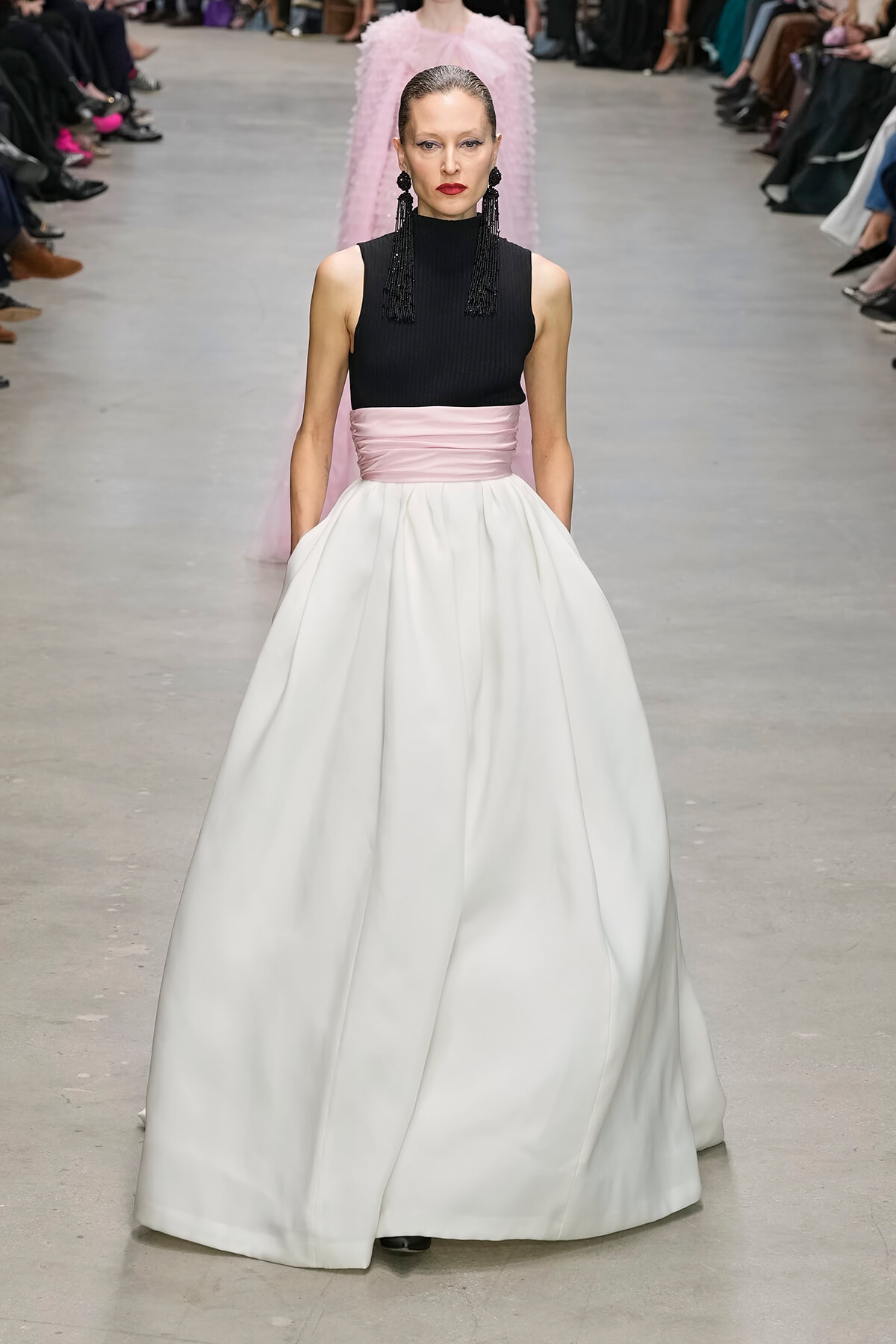 Model walks a runway in a black sleeveless top with a pale pink sash and a full white ball gown, bold red lips, and long black earrings.