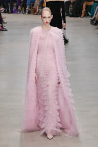 Model walks the runway in a pale pink, multi-layered ruffle gown with a sheer bow at the neckline; audience seated in the background.