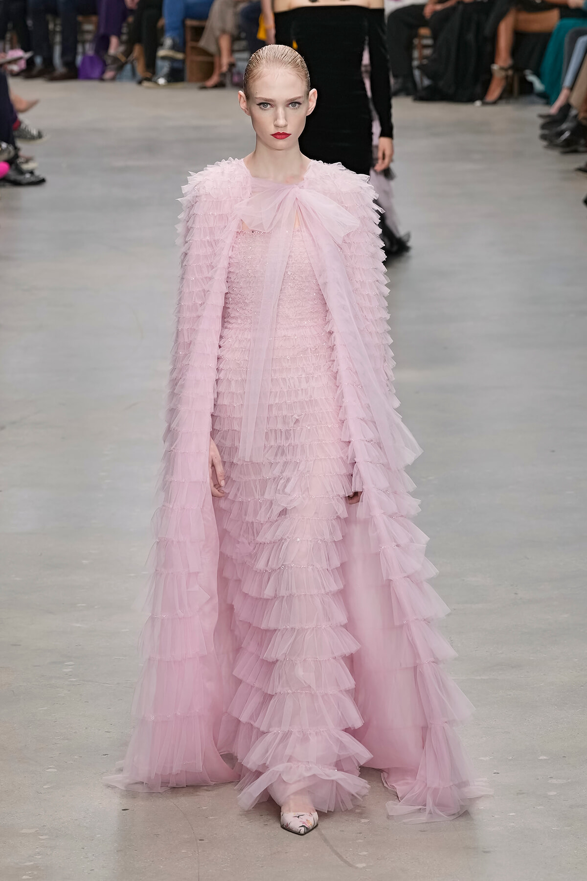 Model walks the runway in a pale pink, multi-layered ruffle gown with a sheer bow at the neckline; audience seated in the background.
