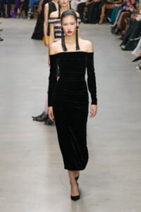 Model walking the runway in an off-the-shoulder black velvet midi dress with long tassel earrings and red lipstick.