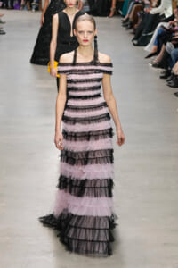 Model walks a runway in an off-the-shoulder pink and black tiered tulle gown with long black fringe earrings, audience in background.