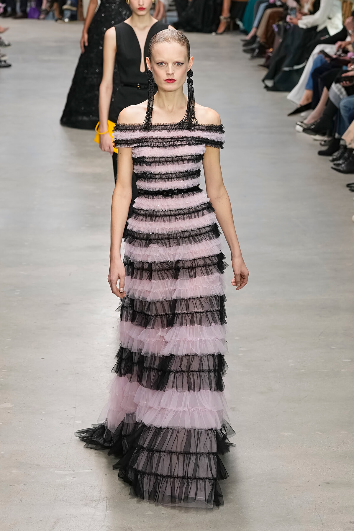 Model walks a runway in an off-the-shoulder pink and black tiered tulle gown with long black fringe earrings, audience in background.
