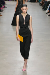 Model wearing a black sleeveless dress with a V keyhole neckline, belt, and yellow clutch on a runway.