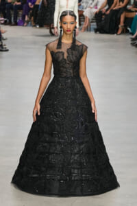 Model walks a runway in a floor-length black gown with sheer neckline and intricate beadwork, accessorized with long black earrings.