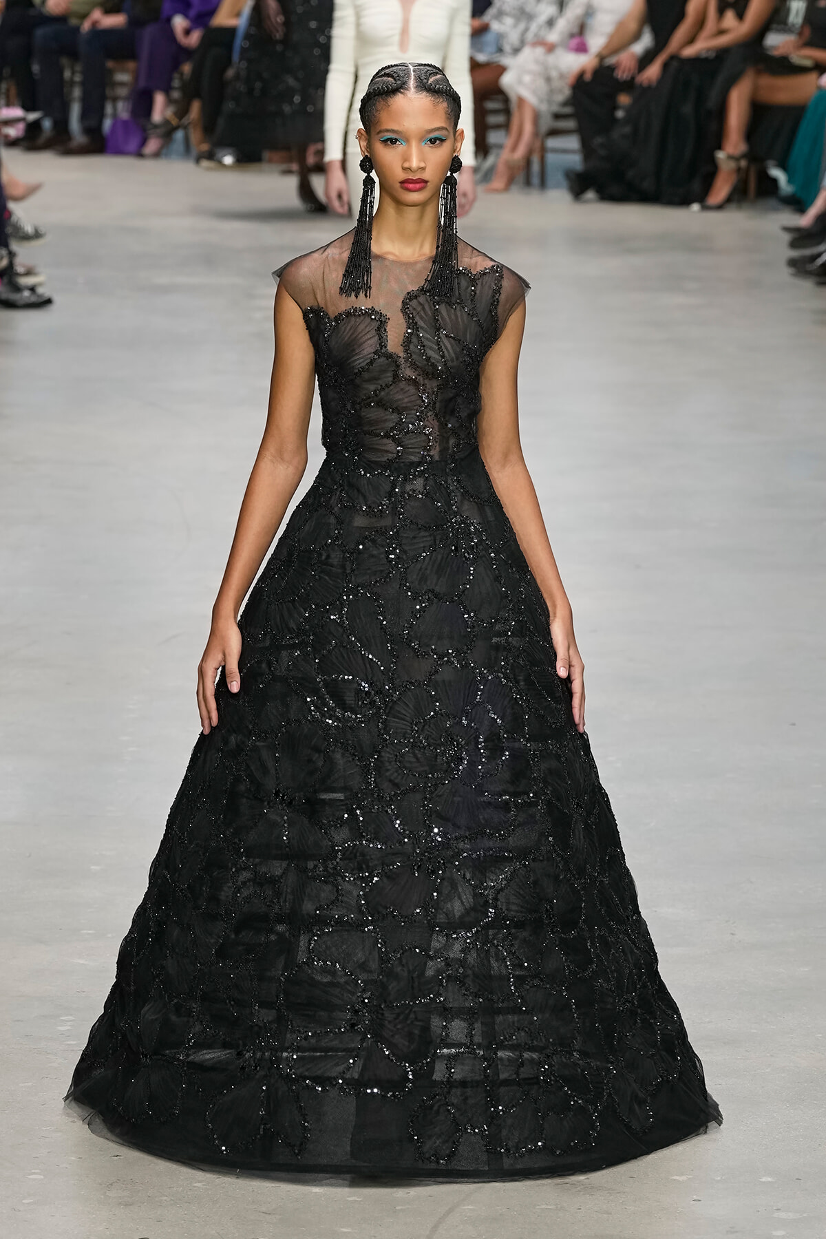 Model walks a runway in a floor-length black gown with sheer neckline and intricate beadwork, accessorized with long black earrings.