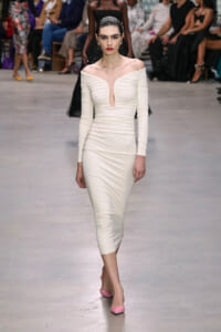Model walks a runway in an off-the-shoulder white ruched midi dress with a plunging neckline, paired with pink pumps; audience seated in the background (out of focus).