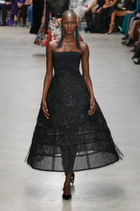 Model walking a runway in a strapless black ball gown with beaded lace overlay and long black fringe earrings, audience in background.