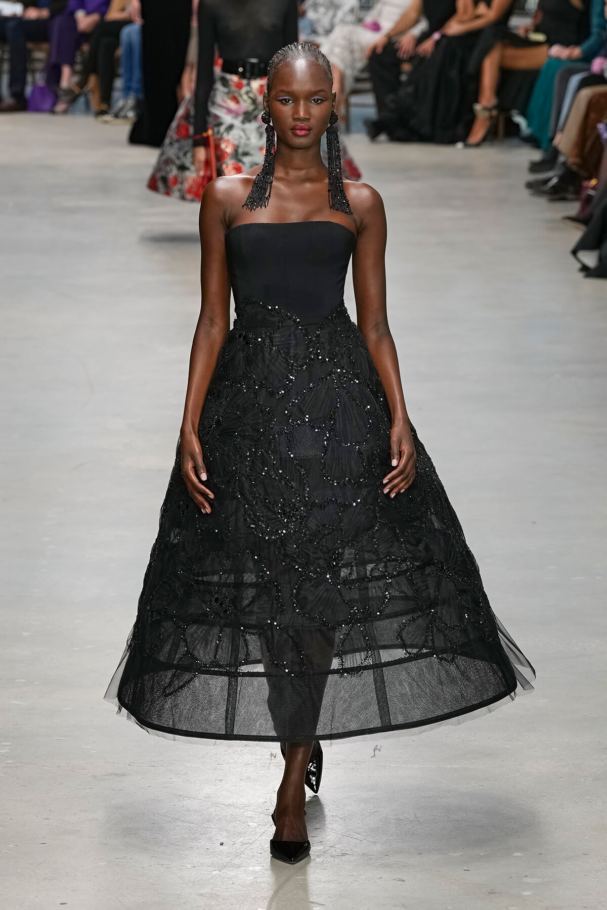 Model walking a runway in a strapless black ball gown with beaded lace overlay and long black fringe earrings, audience in background.