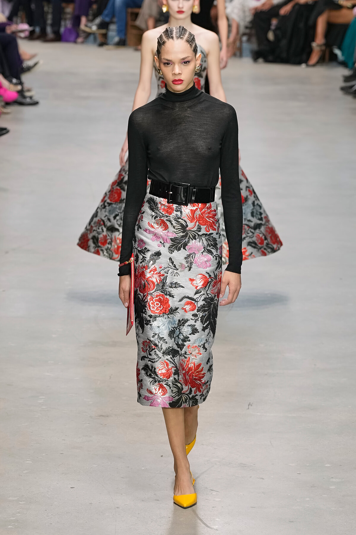 Model walks the runway in a black sheer turtleneck and a gray-red floral midi skirt with a wide black belt.