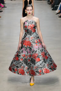Model walks a fashion runway in a strapless, gray floral ball gown with red, pink, and black blooms; bright yellow heels complete the look.