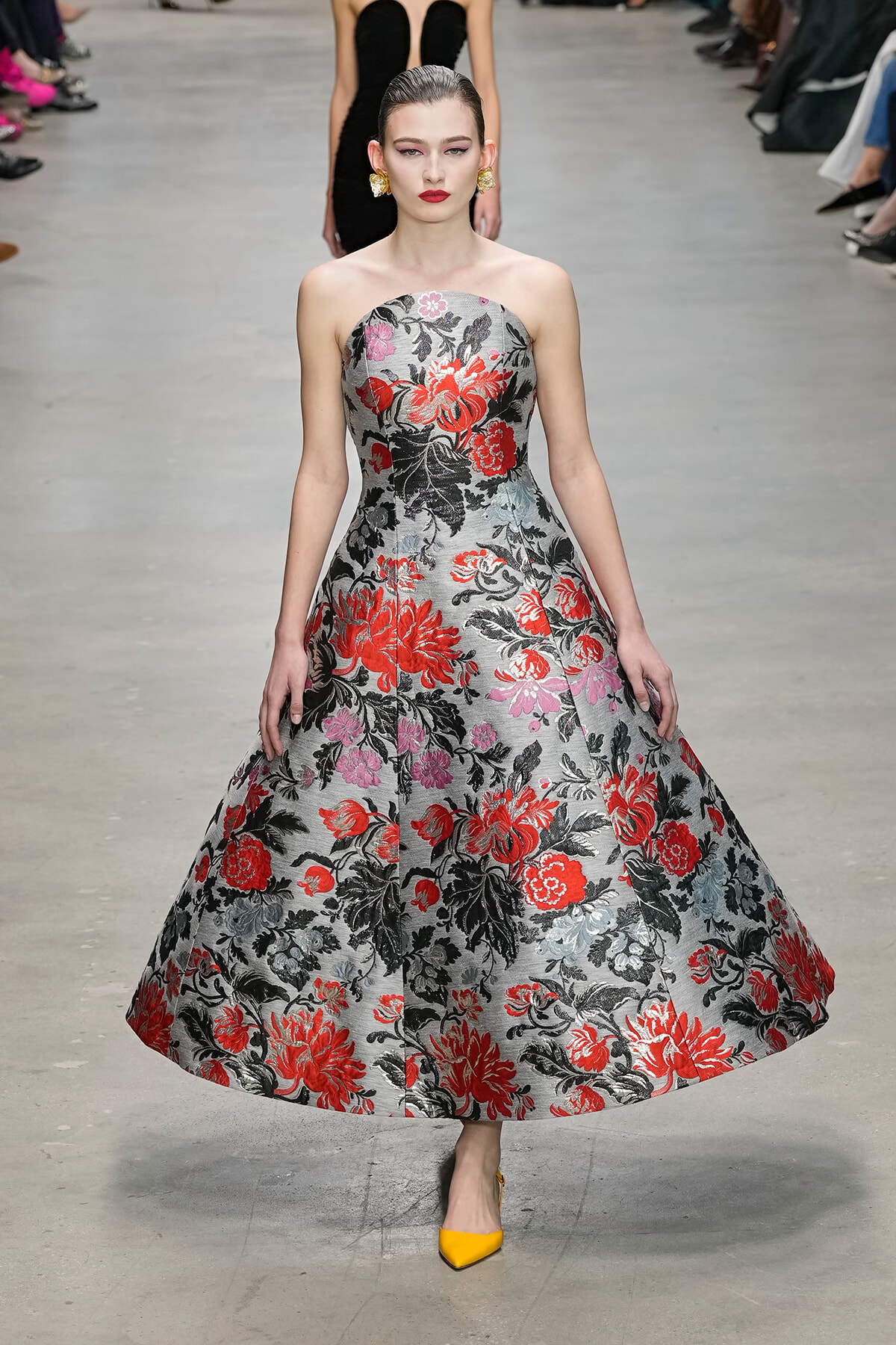 Model walks a fashion runway in a strapless, gray floral ball gown with red, pink, and black blooms; bright yellow heels complete the look.