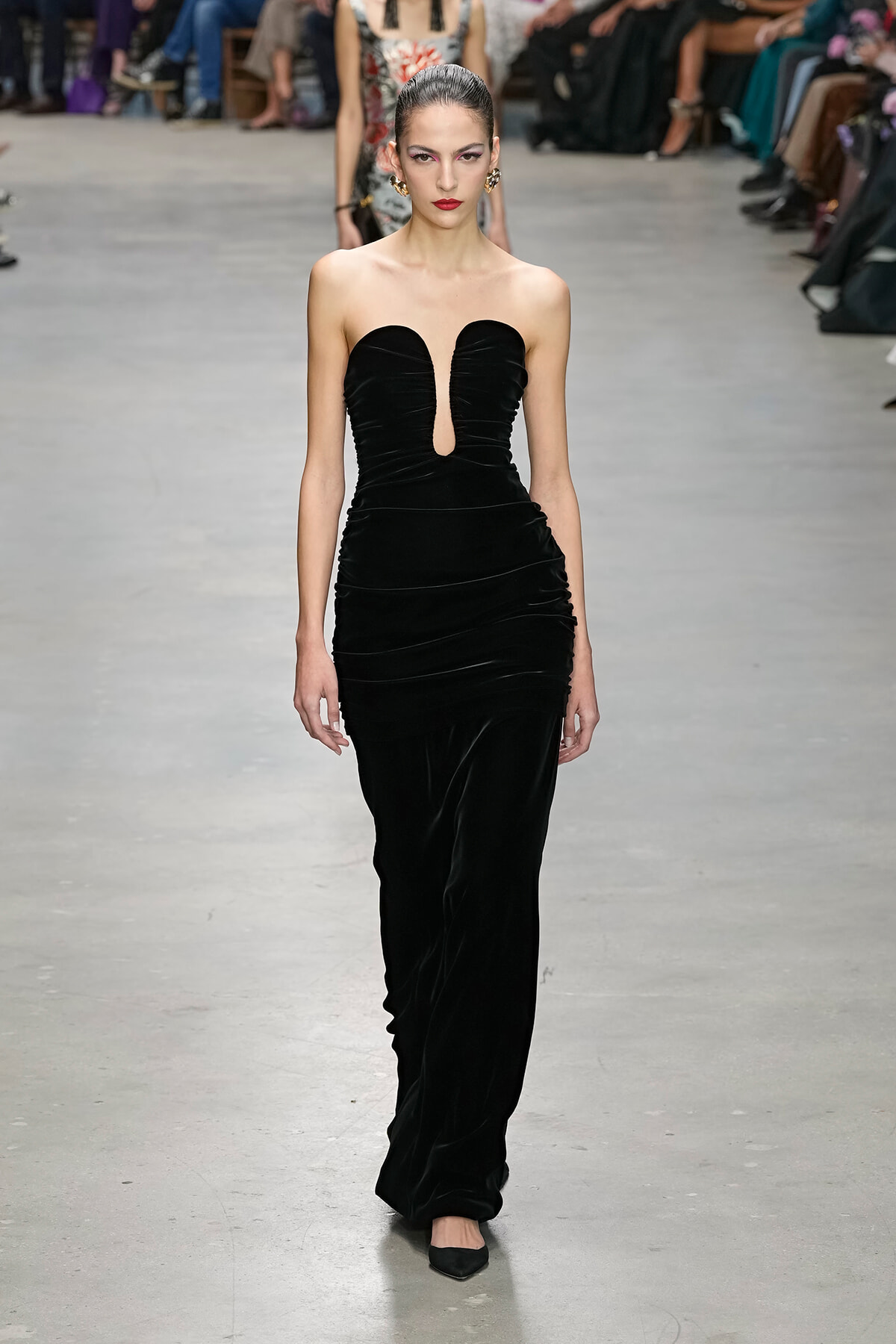Model walking a runway in a strapless black velvet gown with a sculpted notch at the bust.