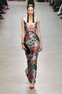 Model on a runway wearing a silver floral gown with red and black flowers, black drop earrings, red heels, and a black clutch.