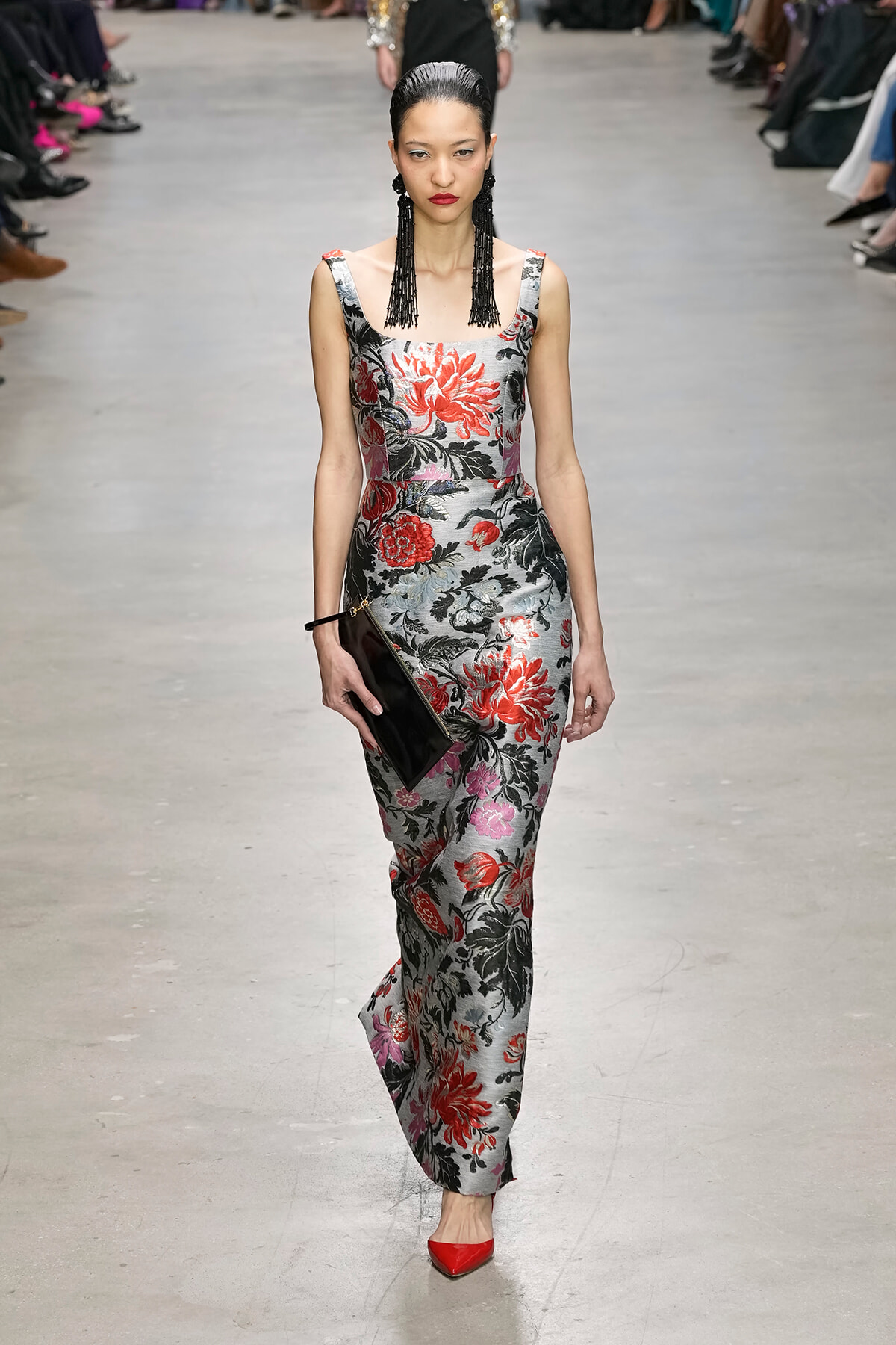 Model on a runway wearing a silver floral gown with red and black flowers, black drop earrings, red heels, and a black clutch.