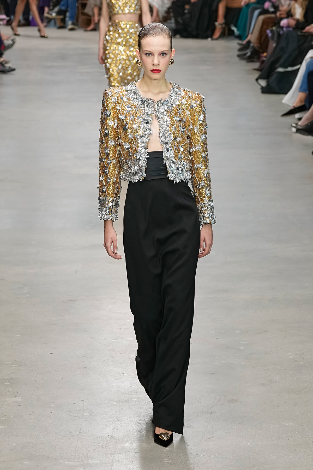 Runway model in a gold and silver sequined jacket with floral embellishments over black wide-leg pants, red lipstick, and a sleek bun.