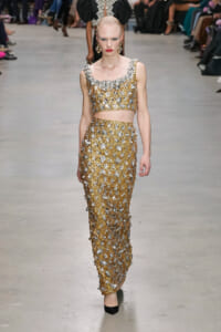 Model walking a fashion show wearing a gold and silver beaded two-piece ensemble with floral appliqués and matching statement earrings, on a neutral runway.