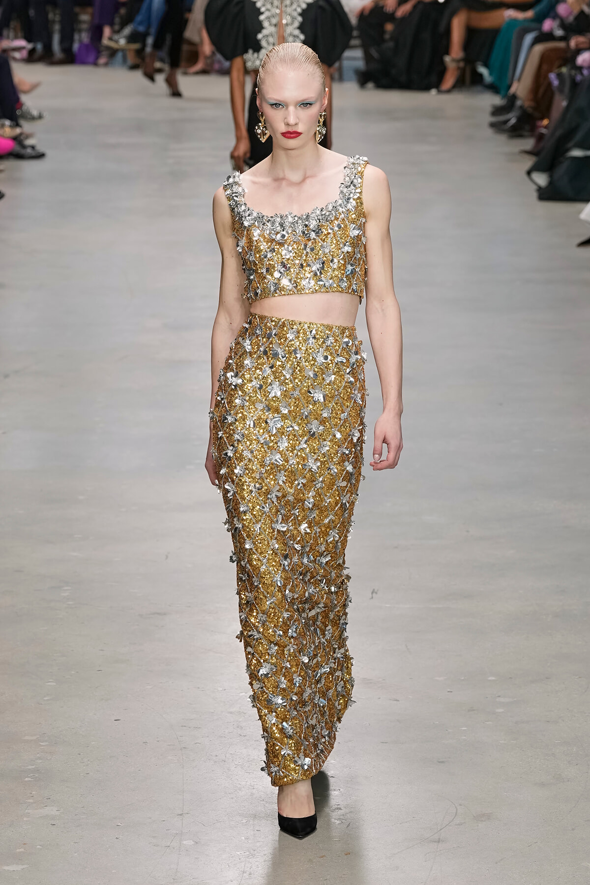 Model walking a fashion show wearing a gold and silver beaded two-piece ensemble with floral appliqués and matching statement earrings, on a neutral runway.