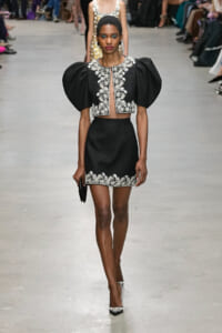 Female model walks a fashion runway in a black two-piece outfit with puff sleeves and white floral embroidery along the neckline and hem.