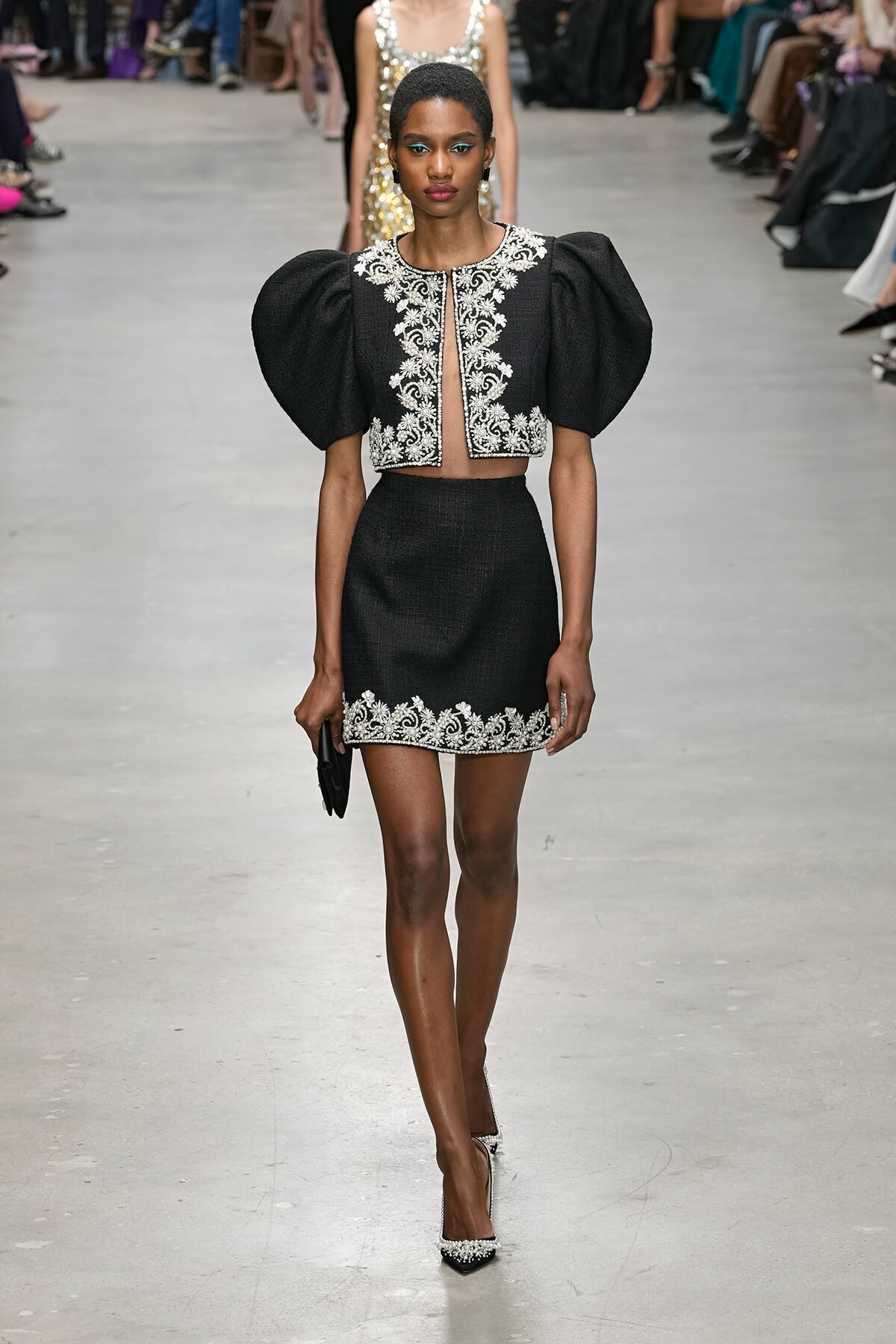 Female model walks a fashion runway in a black two-piece outfit with puff sleeves and white floral embroidery along the neckline and hem.