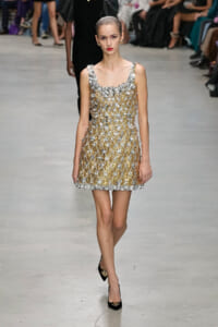 Model walking the runway in a knee-length gold and silver embellished dress with floral appliqués and lattice pattern, black heels.