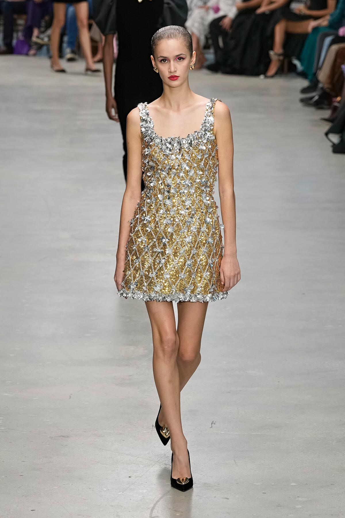 Model walking the runway in a knee-length gold and silver embellished dress with floral appliqués and lattice pattern, black heels.