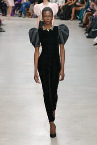 Model walks a fashion runway wearing a black velvet jumpsuit with dramatic puff sleeves and a jeweled collar necklace; audience blurred in background.